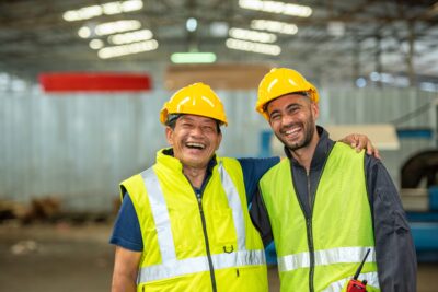 Two happy factory workers wearing hard hats and safety vests standing with arms crossed.